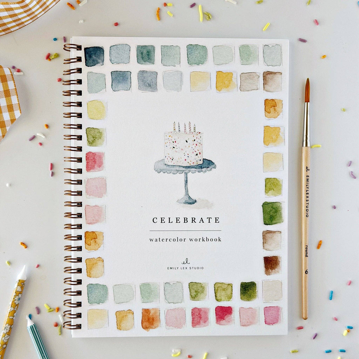 A spiral-bound watercolor workbook titled 'CELEBRATE' with a watercolor cake illustration on the cover, surrounded by a border of painted color swatches. The notebook is photographed from above on a white surface with scattered confetti, alongside a paintbrush and other drawing tools.