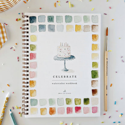 A spiral-bound watercolor workbook titled 'CELEBRATE' with a watercolor cake illustration on the cover, surrounded by a border of painted color swatches. The notebook is photographed from above on a white surface with scattered confetti, alongside a paintbrush and other drawing tools.