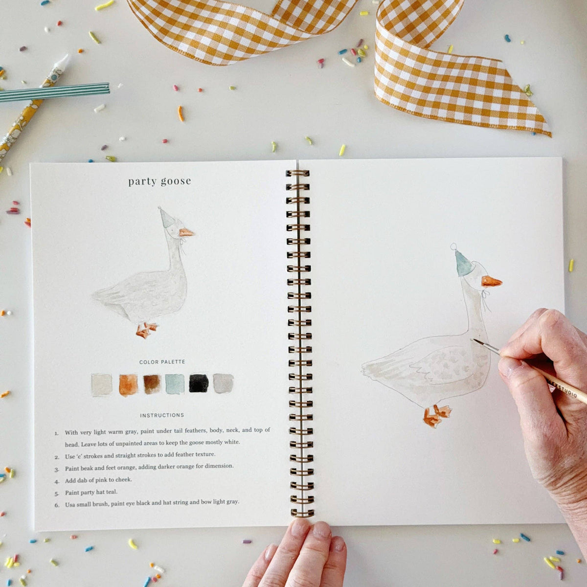 Open book with illustrations of a goose and color palette, surrounded by decorative ribbons and pens.