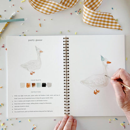 Open book with illustrations of a goose and color palette, surrounded by decorative ribbons and pens.