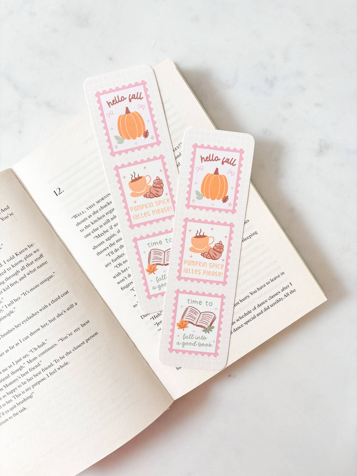 Two decorative bookmarks with a fall theme, featuring illustrations of pumpkins, acorns, and books with pink borders, resting on an open book over a light marble surface.