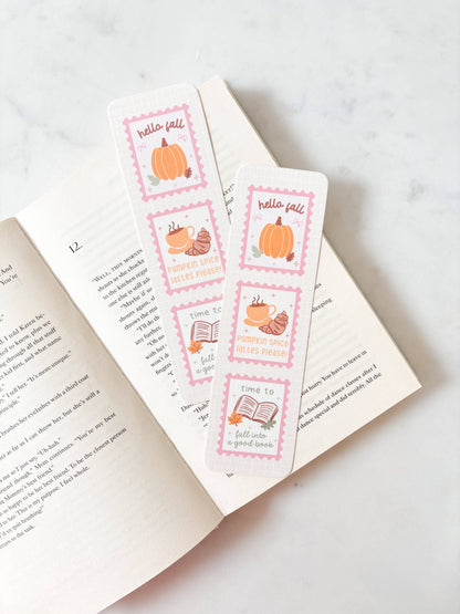 Two decorative bookmarks with a fall theme, featuring illustrations of pumpkins, acorns, and books with pink borders, resting on an open book over a light marble surface.