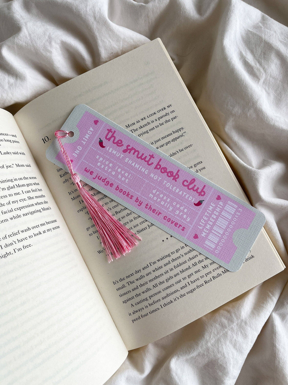A pink-and-light-blue bookmark shaped like a book ticket, with "the smut book club" on it and a pink tassel, resting on an open book on white fabric.
