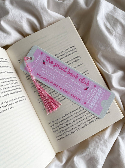 A pink-and-light-blue bookmark shaped like a book ticket, with "the smut book club" on it and a pink tassel, resting on an open book on white fabric.