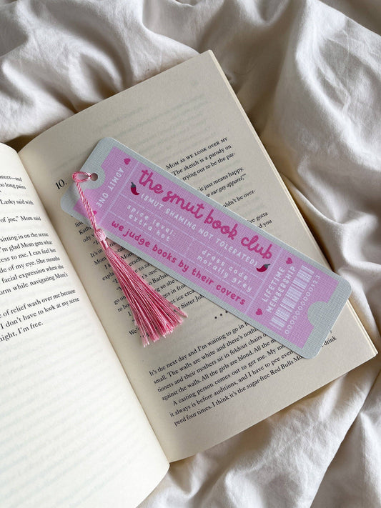 A pink-and-light-blue bookmark shaped like a book ticket, with "the smut book club" on it and a pink tassel, resting on an open book on white fabric.