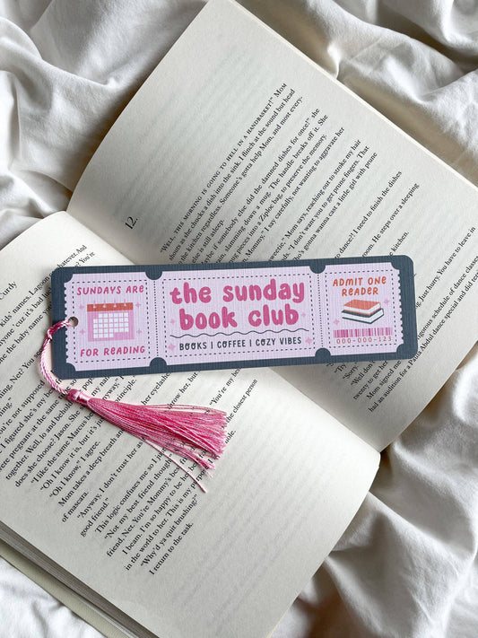 A rectangular paper bookmark with a pink tassel lies across an open book on white fabric. The bookmark has a dark gray border and reads 'the sunday book club' with small ticket-style illustrations on each side.